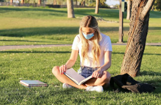 Young masked woman college student reading textbook outdoors.