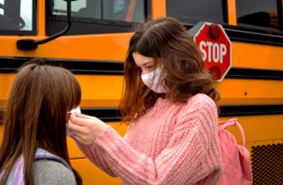 Young sisters help each other put on face masks before boarding school bus.