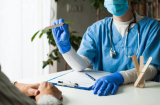 Doctor at desk in face mask and gloves with tongue depressor; patient hands visible in foreground
