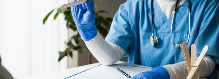 Doctor at desk in face mask and gloves with tongue depressor; patient hands visible in foreground