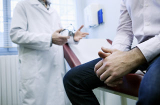 Man sits in foreground on table in doctor's office, only visible from mid-chest to knees, with doctor in background, hands out as though emphasizing a discussion point