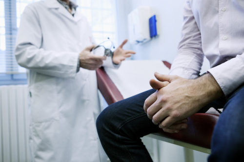 Man sits in foreground on table in doctor's office, only visible from mid-chest to knees, with doctor in background, hands out as though emphasizing a discussion point