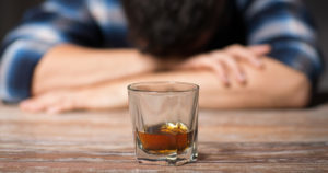 man with his head down on a desk behind an almost empty glass of alcohol