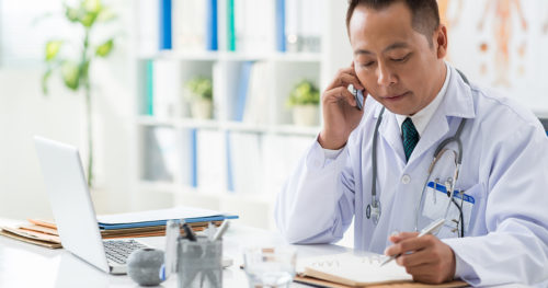 Male Asian doctor at desk speaking on phone with notepad and laptop in front of him
