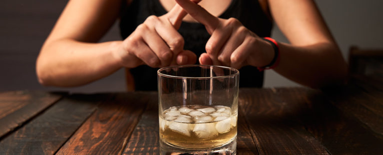 woman behind an glass of alcohol with ice, fingers crossed as if to say "no"