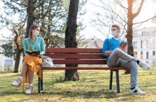 Two people with masks sit far apart from each other on a bench to help prevent the spread of COVID.