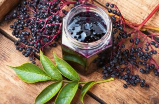 elderberries displayed on a cutting board with a jar of elderberry jam in the center of the image and a sprig of leaves in the lower left corner