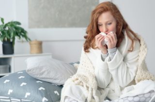 Sick woman sitting on bed with blanket over shoulders as she blows her nose into a facial tissue