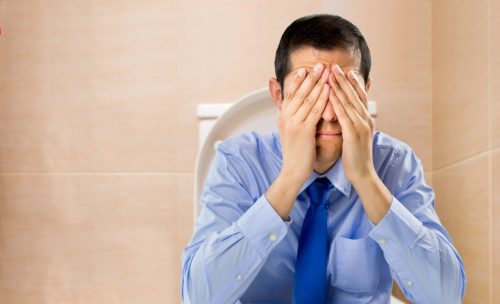 Man in light blue shirt and dark blue tie sits on toilet with hands over eyes