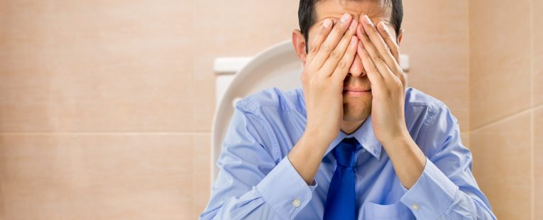 Man in light blue shirt and dark blue tie sits on toilet with hands over eyes